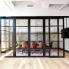 A modern office space featuring a conference room enclosed by large, black-framed glass sliding doors. The room is well-lit, with natural light streaming in from the left side and overhead track lighting illuminating the interior. The conference room contains a wooden table and stylish wooden chairs on a decorative rug.