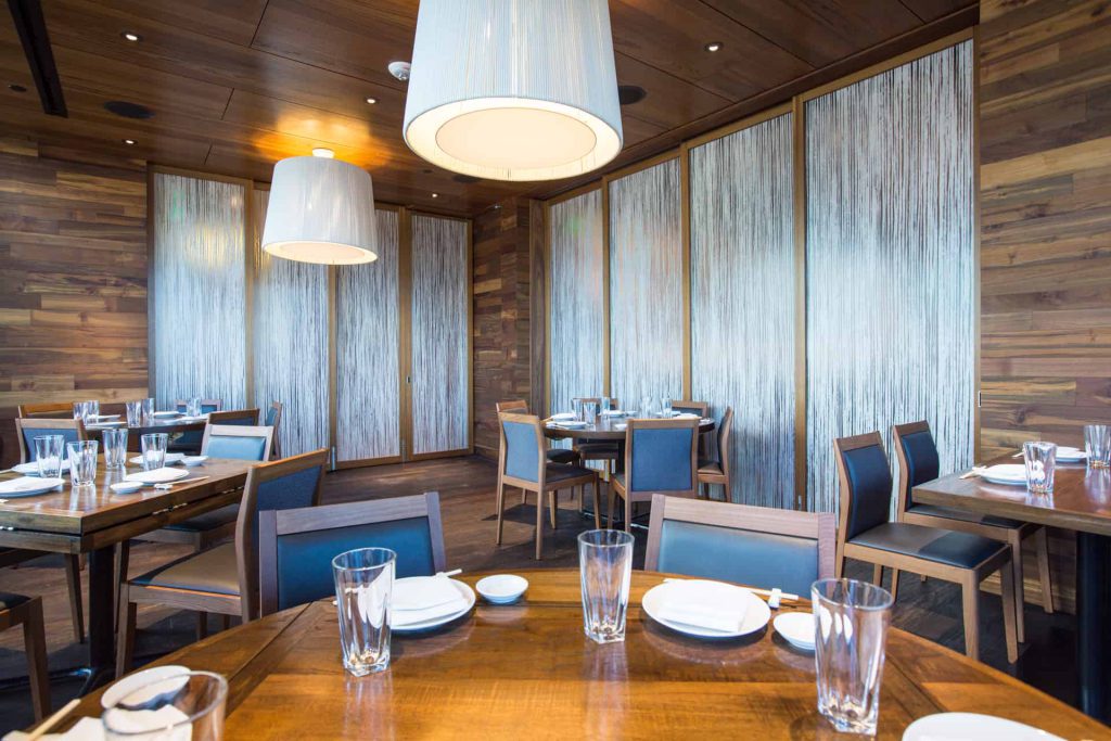 A modern dining area featuring tables and chairs arranged around a series of large sliding wall panels with a unique vertical stripe pattern. The space is well-lit with large, contemporary pendant lights and wooden walls, creating a warm and inviting atmosphere. 