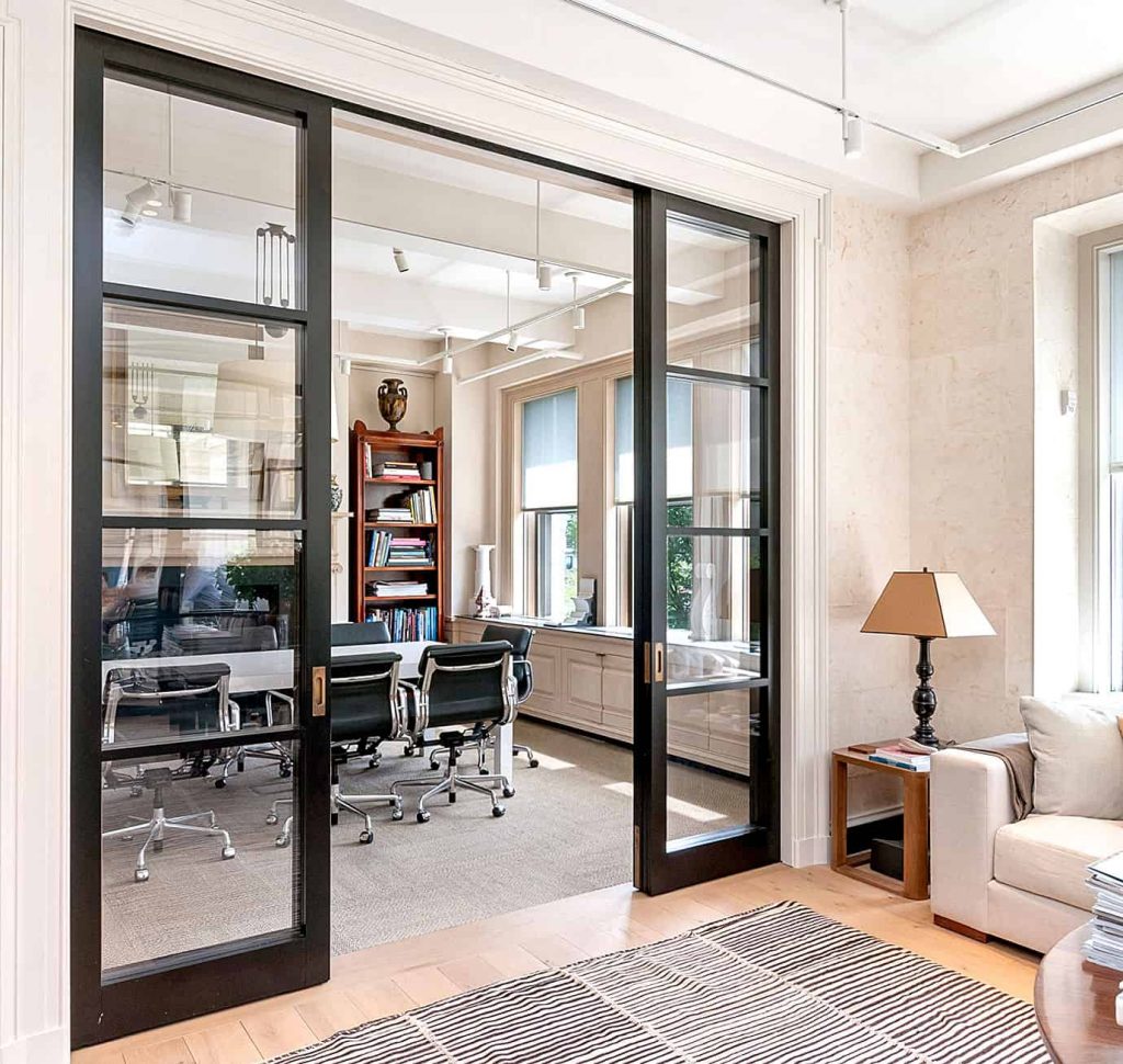 Stylish office space with a conference room separated by black-framed glass sliding doors. Inside, there are ergonomic chairs around a table, bookshelves filled with decor and books, and large windows letting in ample natural light. A cozy seating area with a lamp and side table is visible in the foreground, giving the space a welcoming and sophisticated look.