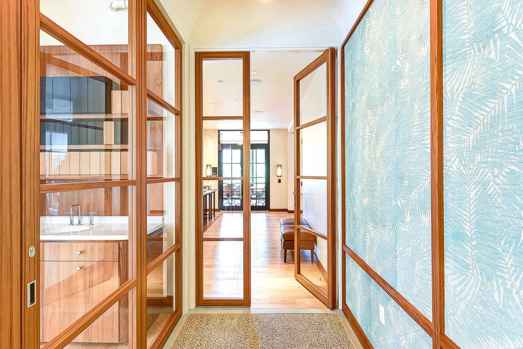 A wooden-framed glass door with multiple clear glass lights in a grid pattern, partially open to reveal a corridor with matching wood-framed doors and warm lighting. The door adds elegance and transparency to the interior, enhancing the open feel of the hallway