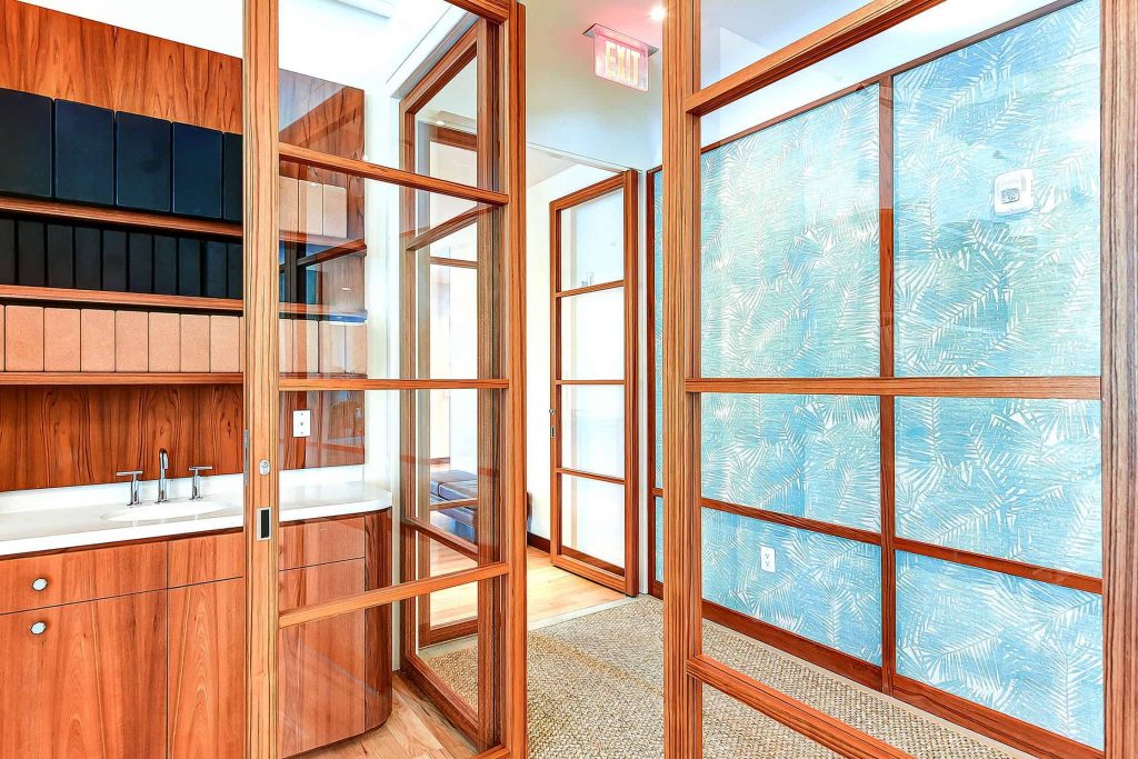 Interior space with wooden-framed glass partitions featuring a grid pattern, dividing areas while maintaining visibility. The room has a sink and cabinetry with natural wood tones, complemented by a light blue accent wall with a subtle leaf pattern, creating a warm and inviting atmosphere.