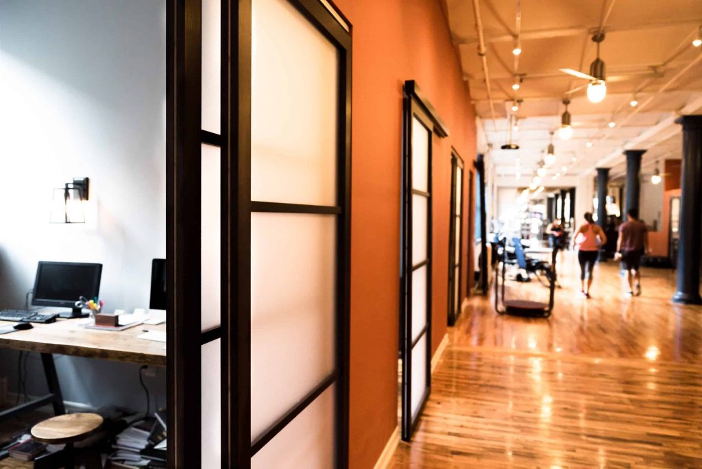A series of sliding doors divides private offices from the rest of a physical therapy clinic. One of the sliding door sets is opened to reveal a well-lit, modern personal office.