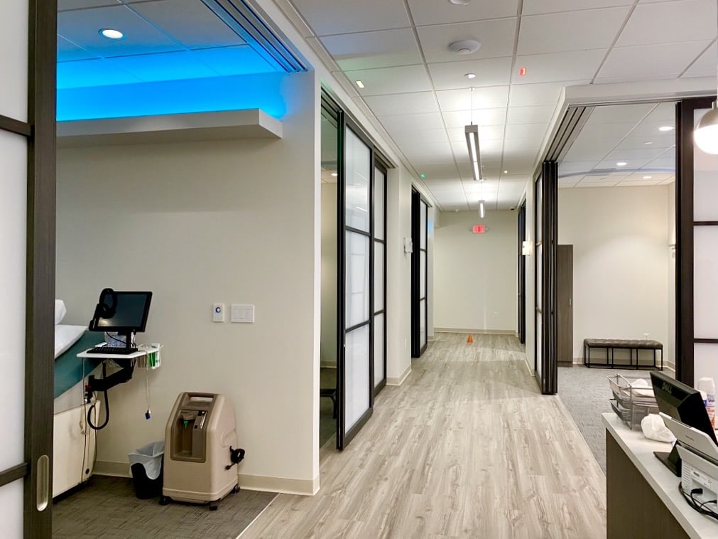 A hallway shot of a brightly lit and modern outpatient facility. Operable door systems flank both sides of the hallway to reveal private treatment areas.