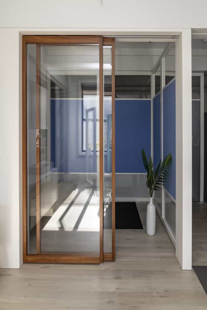 A modern glass sliding door with a wooden frame, featuring clean lines and a sleek design. The door is installed in a bright, minimalist interior with natural light streaming through, creating a warm, inviting atmosphere.