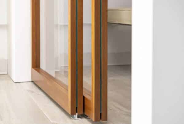 Close-up of the bottom of a wooden-framed glass sliding door, highlighting the craftsmanship of the frame and the smooth sliding mechanism. The door's glass panels reflect light, complementing the light-toned flooring in the modern interior.